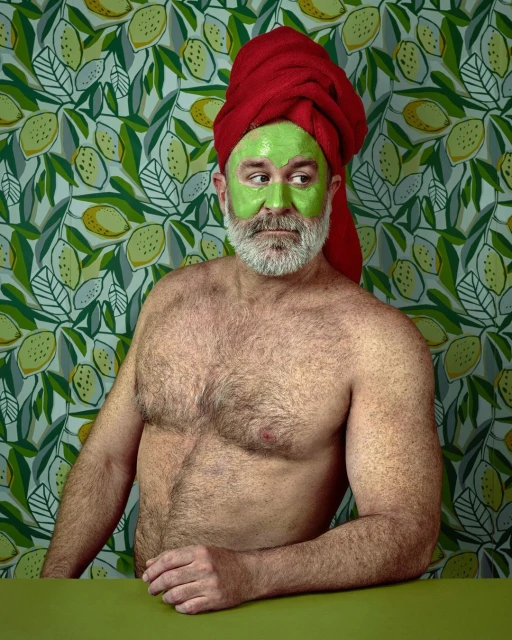 A man with a beard and a red towel on his head, wearing a green facial mask, poses in front of a lemon-patterned wallpaper.